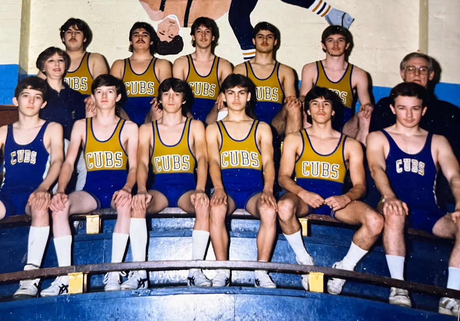 Group of adolescent wrestlers in singlets arranged on three rows of gym bleachers with two adults in the back row; painted gym mural behind them and visible wear on the photo surface.
